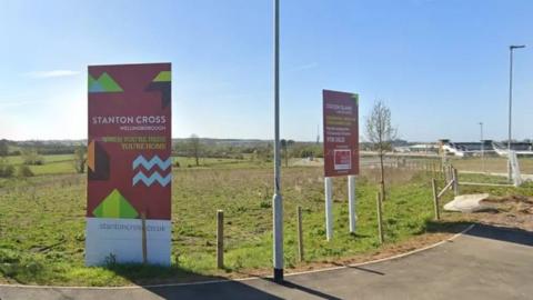 Two large red signs in the ground saying "stanton cross". They are in the grass with fields behind them.
