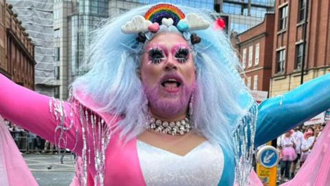 A person with a pink-dyed beard and wearing a pink, blue and white dress with a pink chiffon cape stands with their arms open wide in the Manchester Pride parade