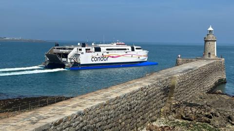 A large white ferry with a Condor logo moves past a small pier and lighthouse.