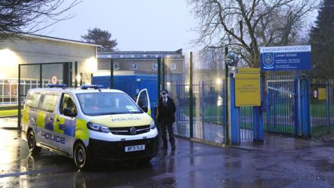A police van is parked inside the gates of Kingsbury High School in Brent at dusk, while an officer stands nearby on a wet road with school buildings and signage visible.