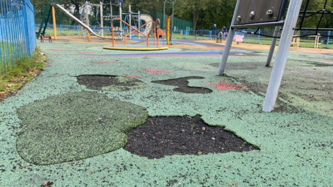 Images show the soft rubber surface on a children's playground broken and missing in places around the bottom of some of the equipment