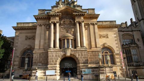 An image of a cream coloured sandstone Edwardian building,  with arched leaded light windows, and arched doorway with Corinthian columns decorative stonework