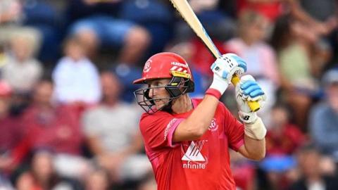 Joe Root, wearing a red shirt and helmet, holds his bat high to his left