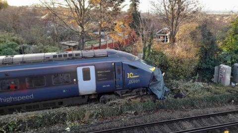 A blue train with the words high speed written on the side in grey. The train has left the track and is in green undergrowth. Trees are in the background