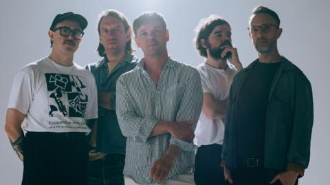 Five men looking into the camera, with light beaming down behind them. Two of the group are wearing glasses and all of them wear serious expressions on their faces. 