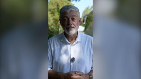 A man with short grey hair, beard and moustache is standing in a shady spot. He is wearing a white shirt.