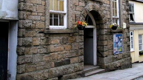 A brown brick building with a large arched door. There are large windows either side of the door. There are flowers in baskets.