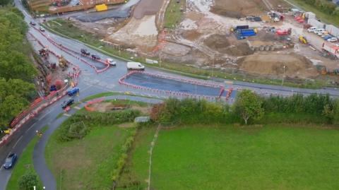 A drone view of the Botley by-pass works with a road running left to right with lines of cones down the middle, a green field in the foreground, and a large area of building site on the other side of the road. It has piles of earth and heavy machinery and a line of cars and vans to the right