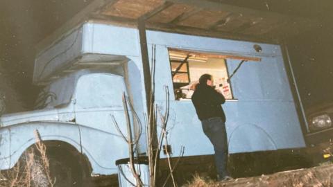 A man in a black jacket stands in front of a pale blue food truck