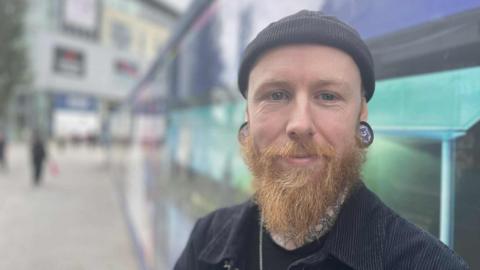 A bearded man in a beanie hat stood near the Eagle Market redevlopment hoardings