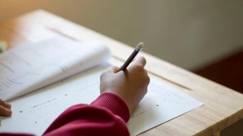 A child's hand on a school desk. The child is holding a pencil and writing in what looks like an exam paper