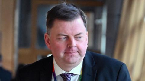 Colin Smyth, a man with flat brown hair, while wearing a suit, white shirt and purple tie and multicoloured lanyard