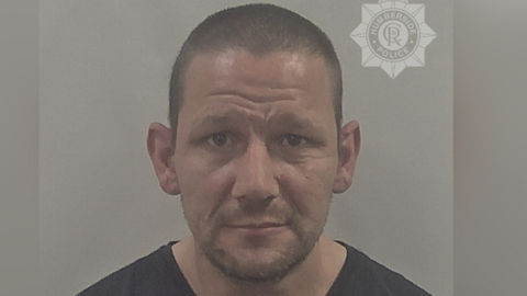 A police custody photo of John Atherton looking at the camera. He has short shaven black hair and short dark stubble on his face. He is wearing a black t-shirt. The wall behind him is grey.