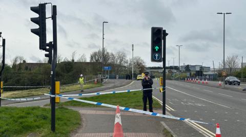 A police officer in uniform mans a cordon next to a busy main road. Lots of police vehicles can be seen parked in the background, and there is lots of blue and white police tape at the entrance of the road. 