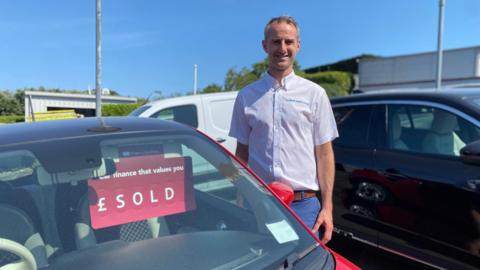 Pete Beausire stands beside a red car displaying a sold sign at a dealership, with other vehicles, a white van, and buildings visible under a clear bright blue sky.