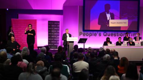 Sadiq Khan, wearing a dark suit and white shirt, stands on stage at a podium. To his right are assembly members sitting behind a white desk-like structure in two rows. Behind them is a screen and lettering that reads 'People's Question Time'. An audience is visible but blurred in the foreground. A sign language interpreter stands to the left of the photo.