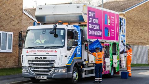 Two bin men in orange hi-vis are pouring bags of waste into a bin lorry with different compartments. A large slogan on the side of the lorry reads "Don't let your recycling go to waste".