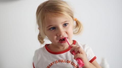 A young girl with blonde hair tied back in pigtails brushes her teeth. She is wearing a white t-shirt which says My heart is full of colours