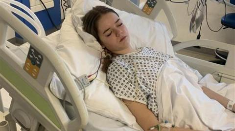 Sophia Speirs, lying in a hospital bed, with her eyes closed, with a gown on and a sheet over her, she has tubes in her and machinery around her. She has long hair tied back. 