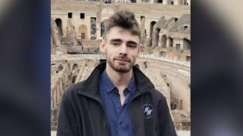 A man with brown hair and stubble looks at the camera