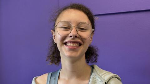 A young woman has brown curly hair tied back in a short ponytail, she is wearing large gold frame glasses and is smiling. She is wearing a grey vest and a grey hoodie. She is sitting in front of a bright purple wall