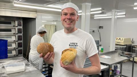 Luke Jenneson stands in his bakery with industrial ovens, mixers and flour covered surfaces in the background. He is wearing white clothing and is holding two bread loaves.