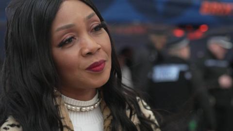Pastor Lorraine Jones Burrell is looking to the right. She has long black hair, wearing a pearl necklace over a white turtle neck top and a animal print jacket. The background is blurred, but shows police officers in black uniforms stood away from her, with their backs to her.
