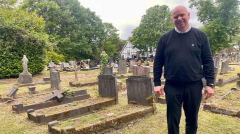 A bald middle-aged man wearing black, standing in a cemetery with graves visible in the background