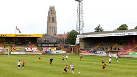 A football match is being played in a small stadium with yellow, black and seating. One team is wearing a yellow and black striped kit, the other is wearing white. The Boston Stump, a church tower, is visible behind the stands. Floodlights are visible in the background and a flags and banners are also hanging in the stands behind the goal.