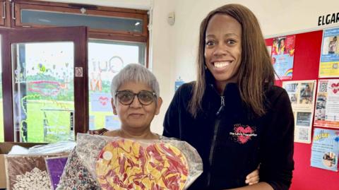 Jagruti Patel and Lorraine Lewis pose for a picture inside a community centre. An open playing field is visible out of double doors behind them. 