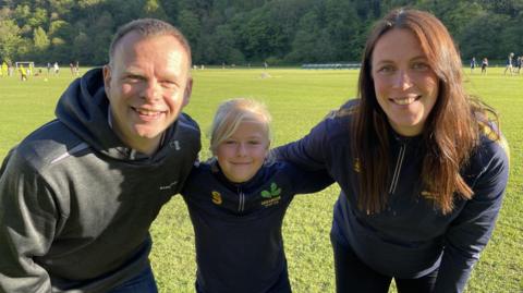 Edie, nine, stands smiling between her dad Matt and her coach Amy Sherratt on the grass at Oakamoor Cricket Club. She wears a navy Oakamoor Cricket Club training top with the club's oak leaf badge. Behind them, junior players take part in a training session against a backdrop of wooded hillside on a bright, sunny evening.