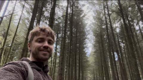 Max Weston in a selfie, smiling down at the camera with tall trees in the background and sky behind them. He is wearing a brown fluffy jumper and has rucksack straps over his shoulders. 