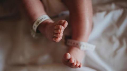 The feet of an unidentifiable newborn baby in a hospital bed. The baby has hospital identification tags on both its ankles - though none of the information is legible.   