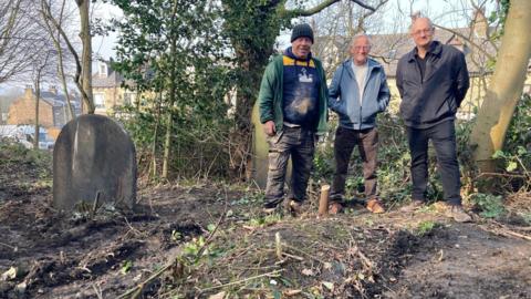 Three men standing at the foot of an unmarked grave with a small tree stump marking the spot
