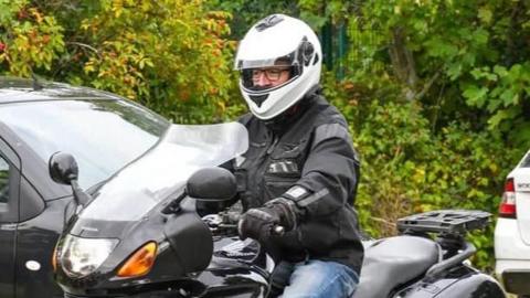 A man wearing a white motorbike helmet and black gloves is seen sat riding a black motorcycle with a wind shield at the front.