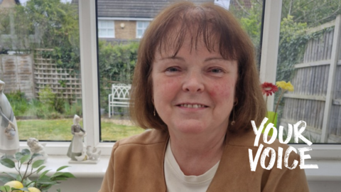 Carole Moore, who is smiling at the camera, is sitting in her conservatory. her back garden can be seen through the windows. 