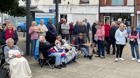 Protesters in Dinnington outside the office of Jake Richards, MP for Rother Valley. Many of them are elderly, and some are sat in camping chairs with blankets covering them.