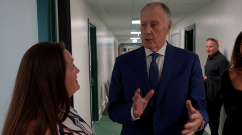 Sir Geoff Hurst gesturing with his hands and talking to the head teacher during a visit to the school. He is wearing a suit and standing in a corridor.