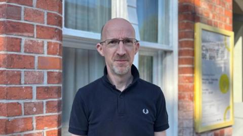 A man wearing glasses smiles slightly at the camera. He is wearing a navy blue polo shirt with the collar turned up. In the background is the sign for Cressington station. 