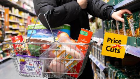 A person wearing a black coat in a supermarket aisle taking pasta off the shelf to put it in their very full basket. A yellow sign which reads reads Price Cuts next to a red pair of scissors hangs fro the shelf.
