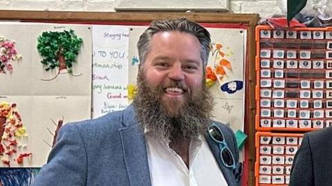 Dan Lentell wearing a blue suit and white shirt, smiling at the camera, in the background is a children's collage on the wall of a classroom.