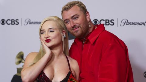 A woman with blonde hair and red lipstick poses with her hand cupping her chin on a red carpet. Standing next to her is Sam Smith who has short hair and a pearl dangling earring. They are wearing a red shirt and has one hand round her waist, with their other holding a small gold trophy.