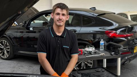 A man with short brown hair and stubble wearing a black polo shirt and orange protective gloves stands beside a black SUV with the bonnet open in a vehicle workshop. A tool trolley holding automotive tools, cleaning cloths and a water bottle is positioned in front of the vehicle. The workshop setting includes additional cars and equipment in the background.