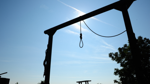 A noose hanging from a gallows. The sun is behind the structure, covering it in shadow.