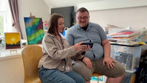 A man in blue top and khaki shirts and glasses looking at mobile phone footage held by a woman in blue jeans and beige top. She has long brown hair