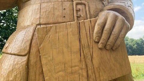 A close up of a wooden statue, showing visible vertical cracks. The statue is a yellow/golden colour and depicts a man holding a book and wearing a rain coat.