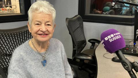 Jacqueline Wilson sat in a radio studio for BBC Radio Sussex. She is wearing a light blue jumper and a big blue necklace, she has grey hair. 