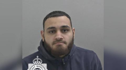 A mugshot of a man with short black hair and a dark beard. He is wearing a black hoodie and looking into the camera.