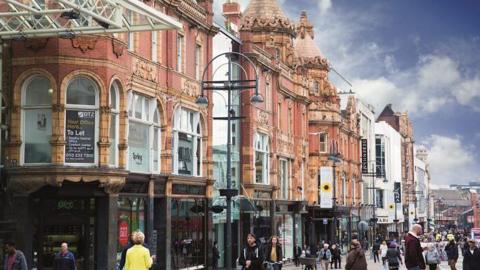 A busy shopping street in Leeds which has a row of Edwardian buildings in terracotta brick with ornate decorations and turretted roofs. 
