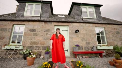A woman in a red dress standing outside a cottage